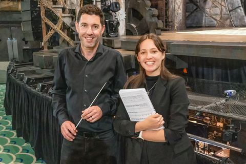 Wicked London Musical Director Ron Crocker, holding a baton, and Assistant Musical Director Millie Davies, holding a music score, beside the orchestra pit at the Apollo Victoria Theatre